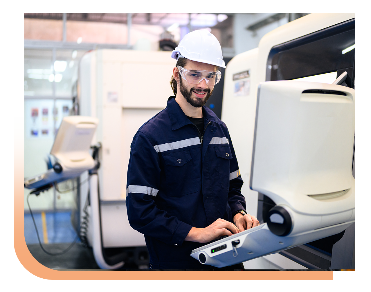 Smiling engineer in a white hard hat and safety glasses operates a control panel on a manufacturing machine in a bright factory.