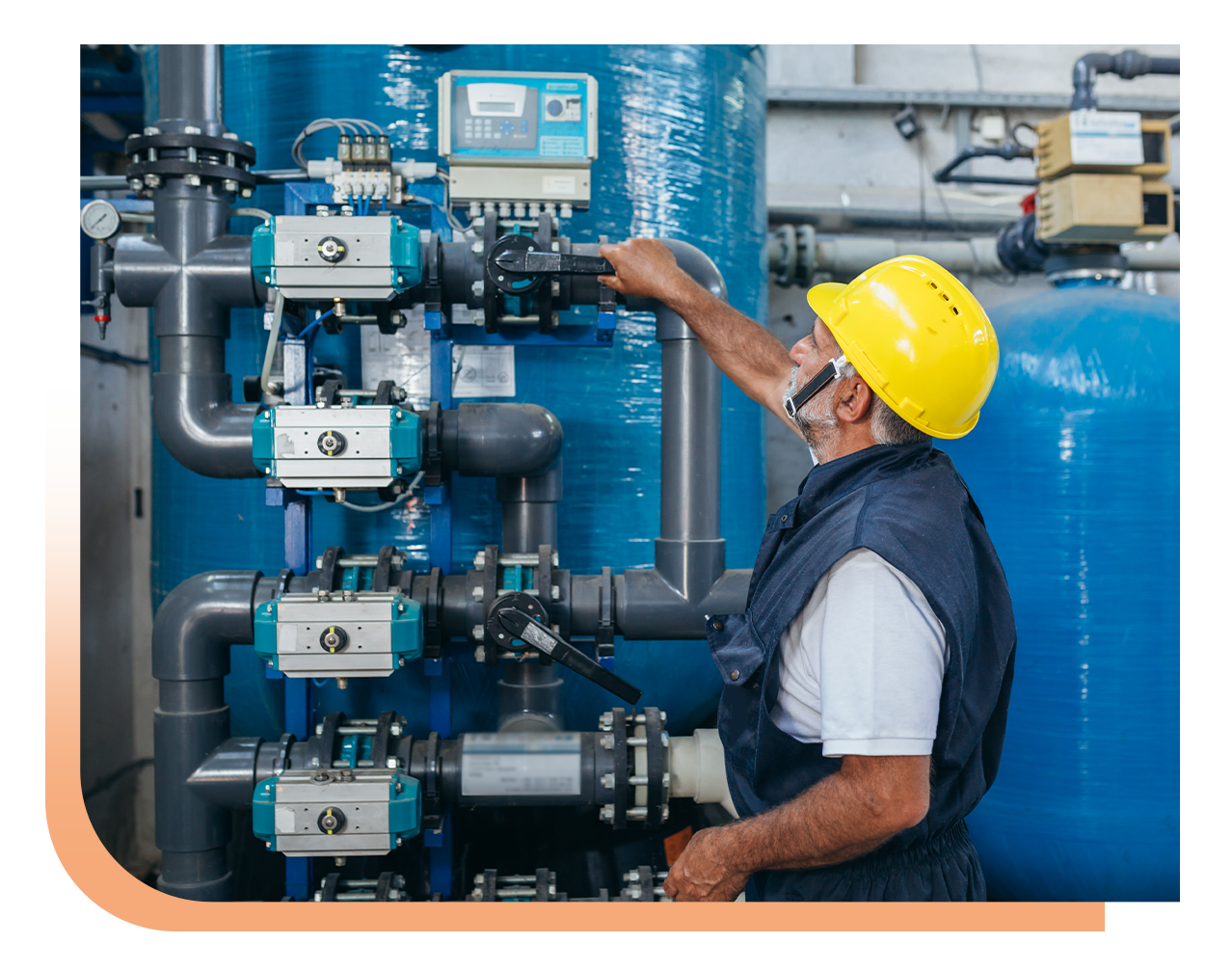 Maintenance worker in a yellow hard hat adjusts valve handles on a complex blue piping system in an industrial plant.