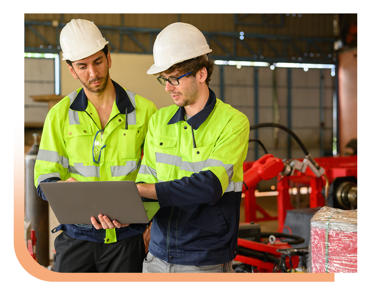 Two construction workers in high-visibility jackets and hard hats examine data on a laptop in an industrial setting, with machinery in the background.