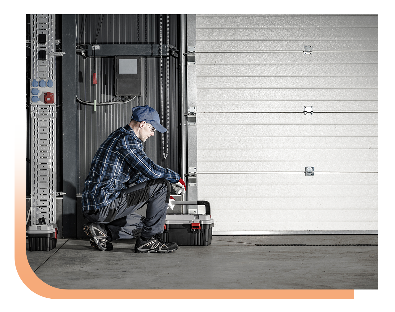 Technician kneels beside an open metal roll-up door in a warehouse, handling a toolbox and red tool case.