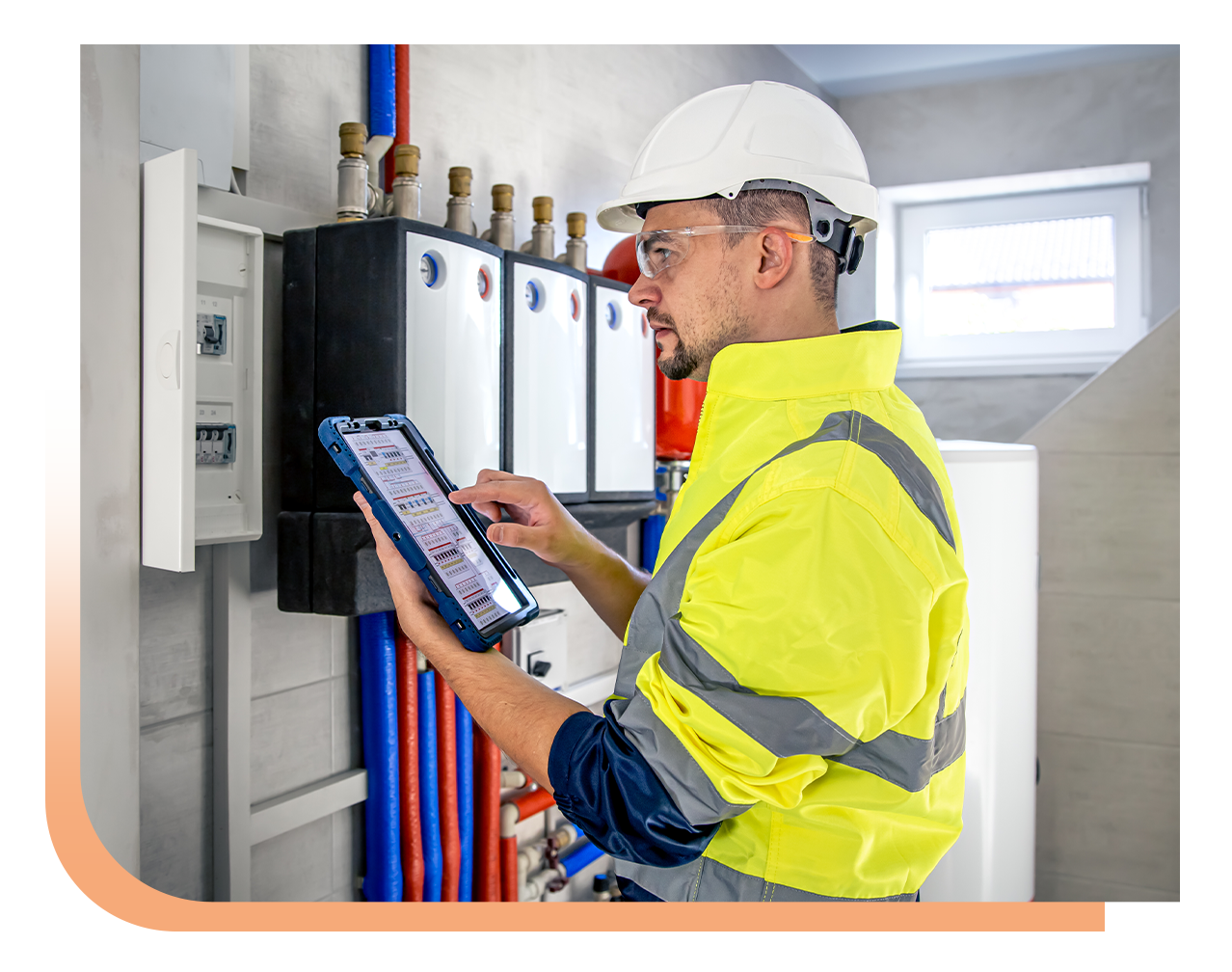 Technician in a high-visibility jacket and hard hat using a tablet to inspect an electrical control panel in a utility room.