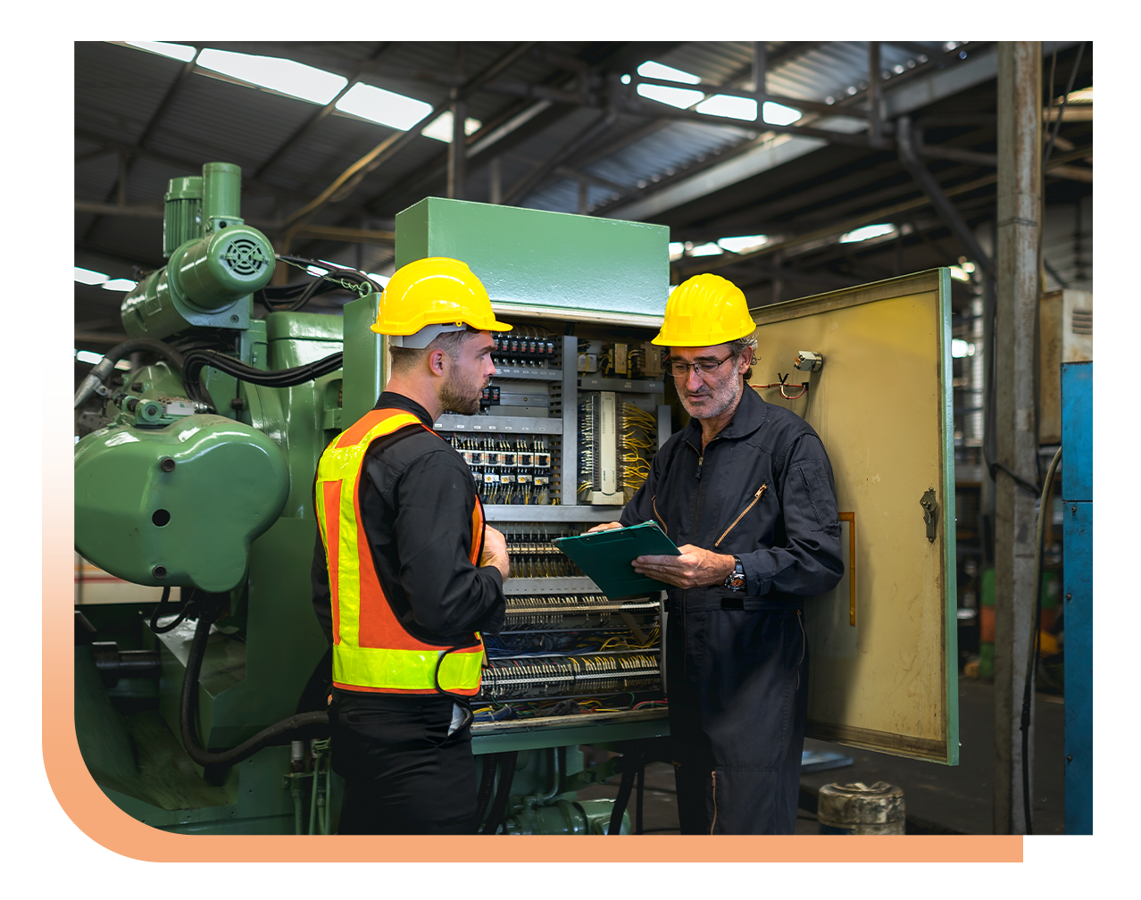 Two male factory workers in yellow hard hats review an open electrical control panel.