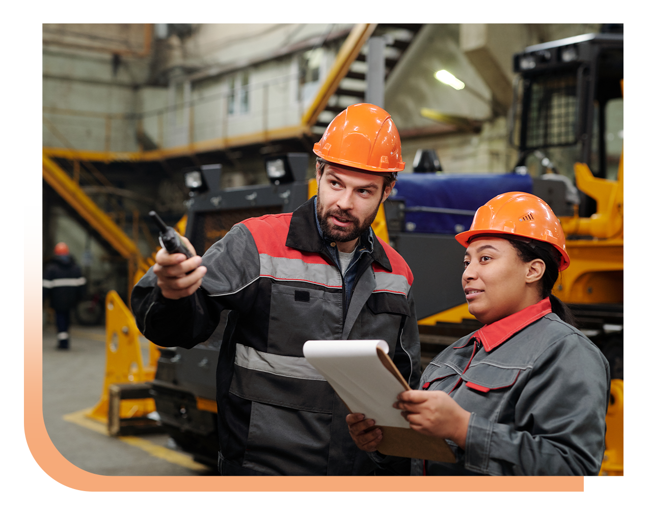 Two factory workers wearing orange hard hats review plans, one points a device while the other holds a clipboard in a busy industrial setting.