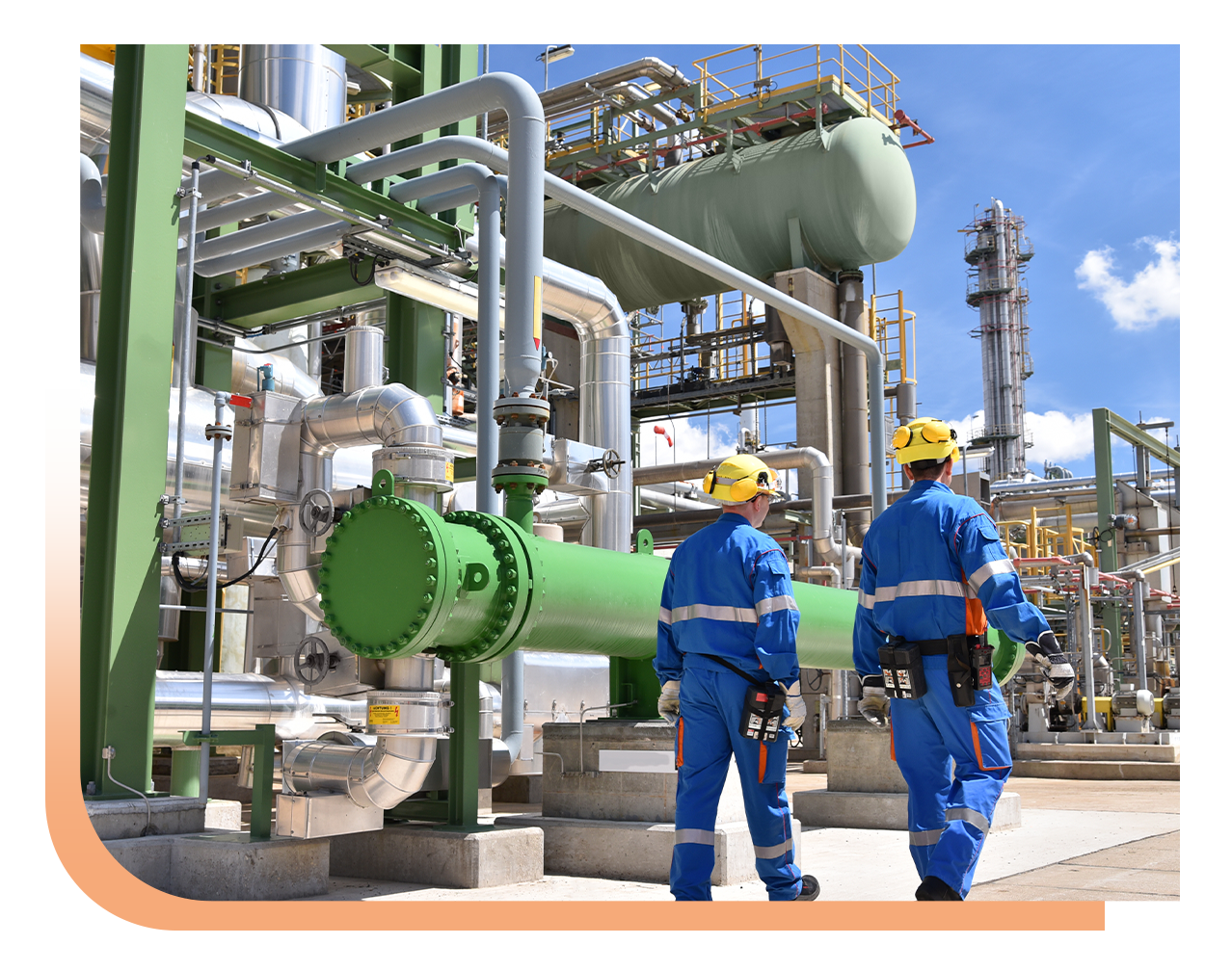 Two workers in blue safety gear and yellow hard hats walk past large green pipes in a complex industrial plant with metal scaffolding and equipment behind them.