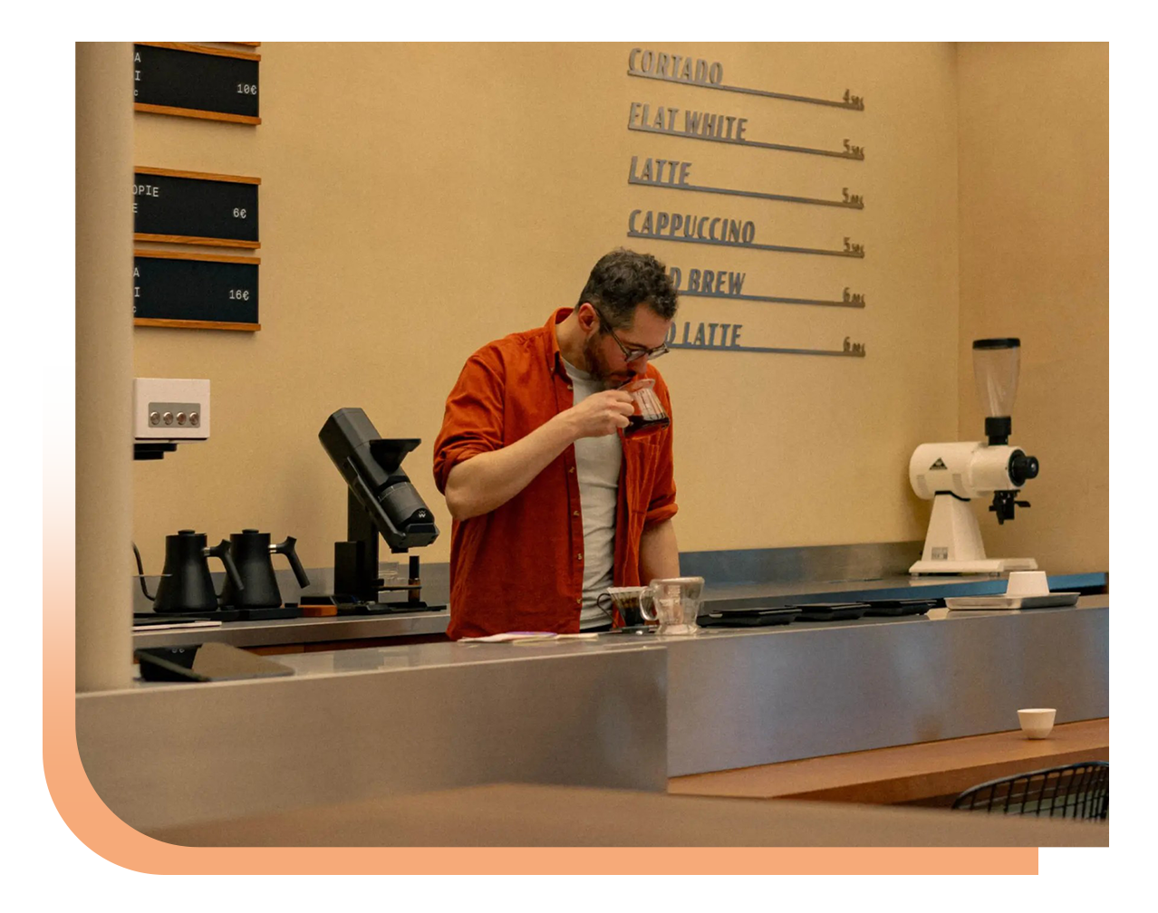Barista in an orange shirt tastes a coffee sample at a cafe counter with espresso gear in the background
