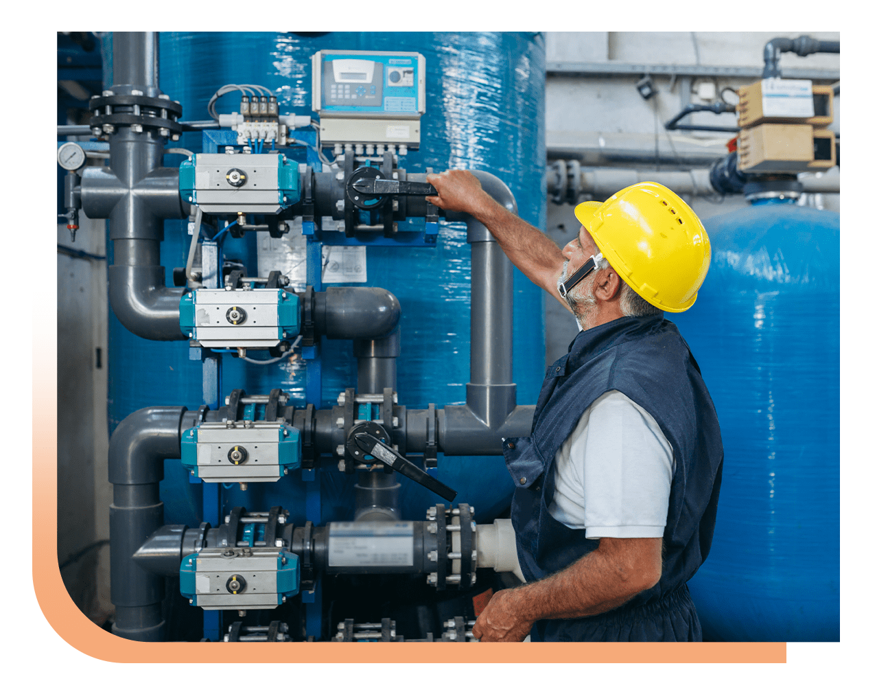 Male technician in a yellow hard hat adjusts industrial valves on a piping system in a facility with blue tanks behind him.