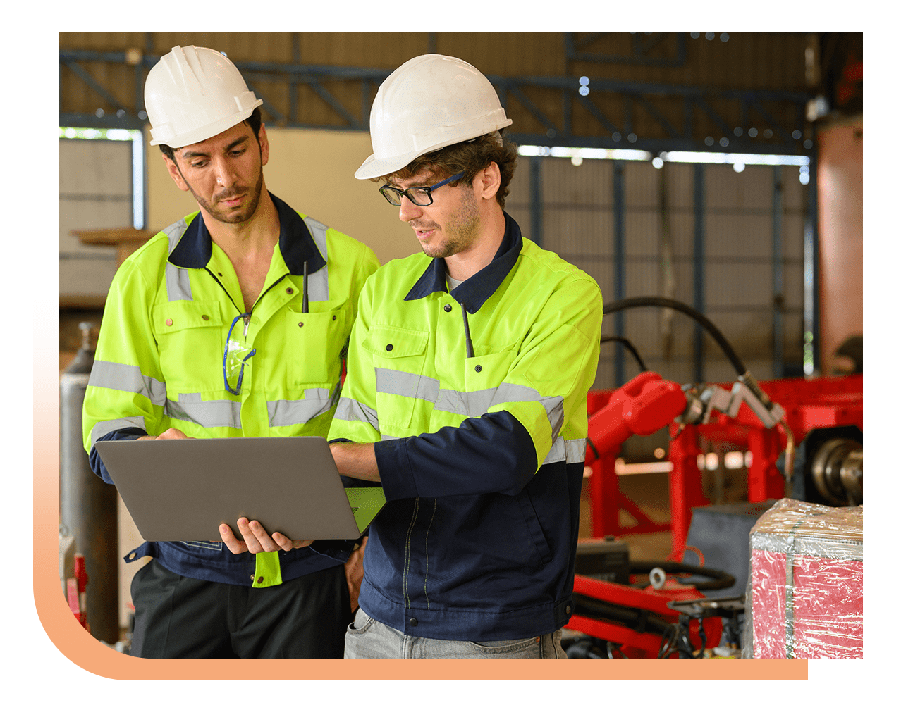 Two construction workers in bright safety jackets and white hard hats review plans on a laptop in a factory setting.