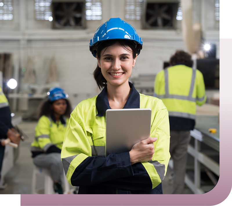 Smiling female engineer in a neon safety jacket and blue hard hat, holding a tablet in a factory setting.