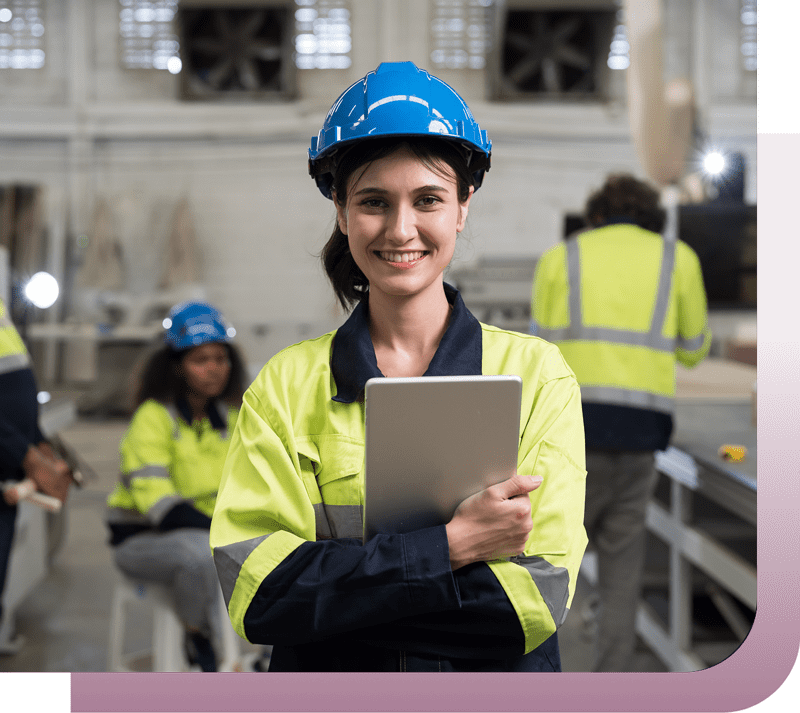 Smiling female worker in a blue hard hat and high-visibility jacket holds a tablet in an industrial setting.